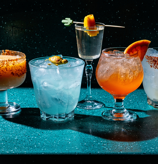 Assorted cocktails on a teal bar, garnished with spices, citrus, and fruit, against a dark background.