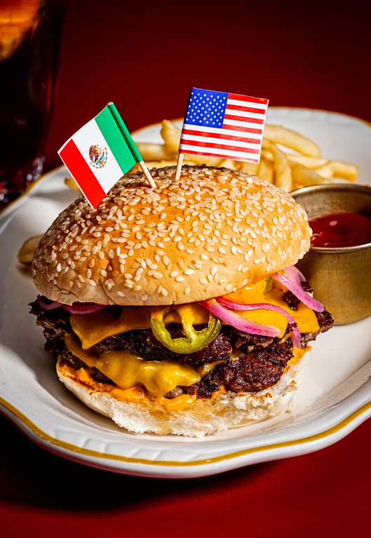 Tall, double cheeseburger with jalapeño, red onion, and sesame seed bun, topped with American and Mexican flags.