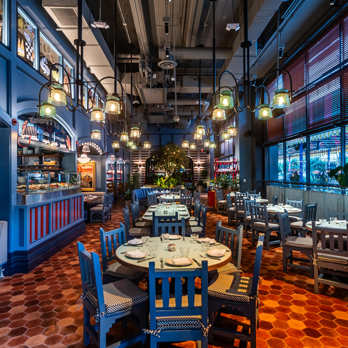 Modern restaurant with blue walls, hexagonal tile floor, hanging lights, and blue and gray chairs. Open kitchen and large windows with ocean views.