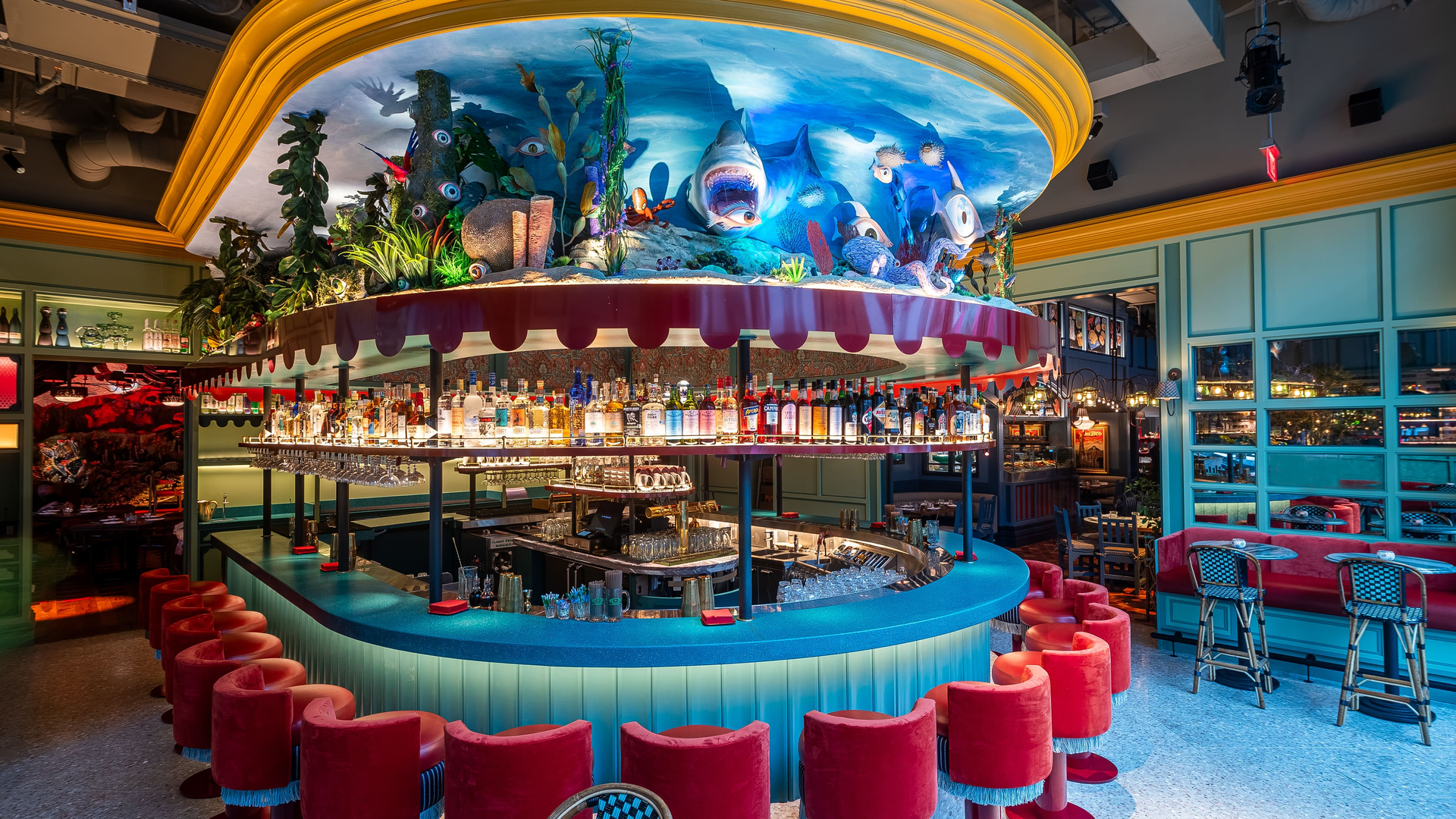 Underwater-themed bar with vibrant decor, red velvet stools, and an array of liquor bottles, illuminated by blue and red lights.