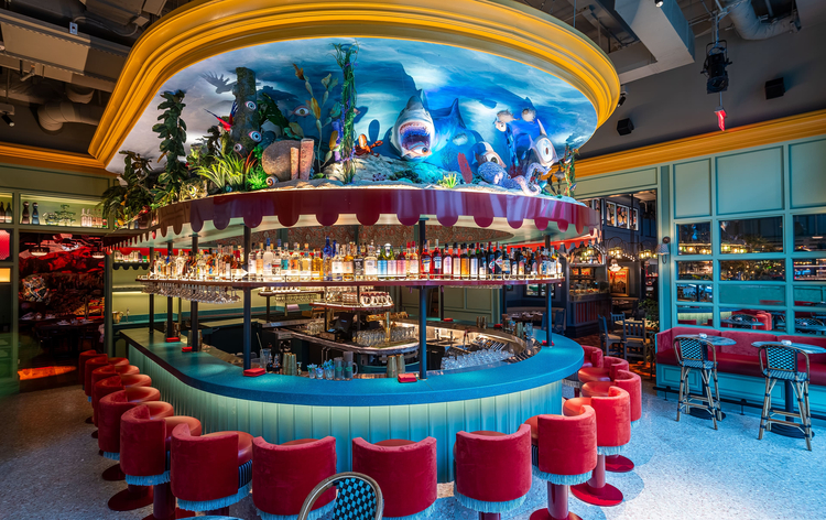 Eclectic bar with vibrant red chairs, blue and green decor, and tank-like structure above.