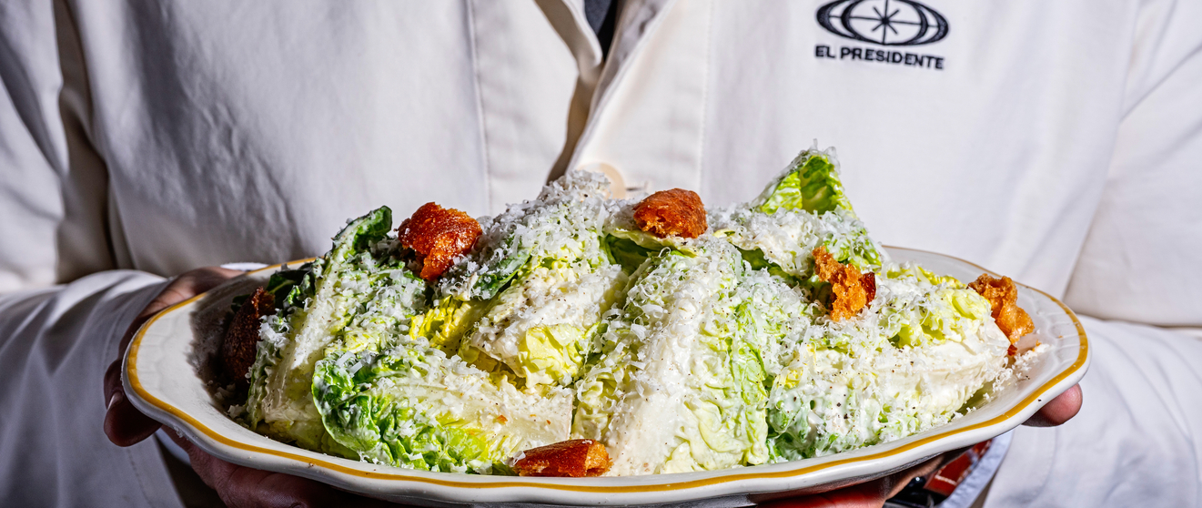 Chef's hand holds a white plate with Caesar salad topped with croutons and grated cheese.
