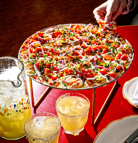A colorful loaded plate of nachos on a raised stand, surrounded by margaritas and lemonade on a red table.