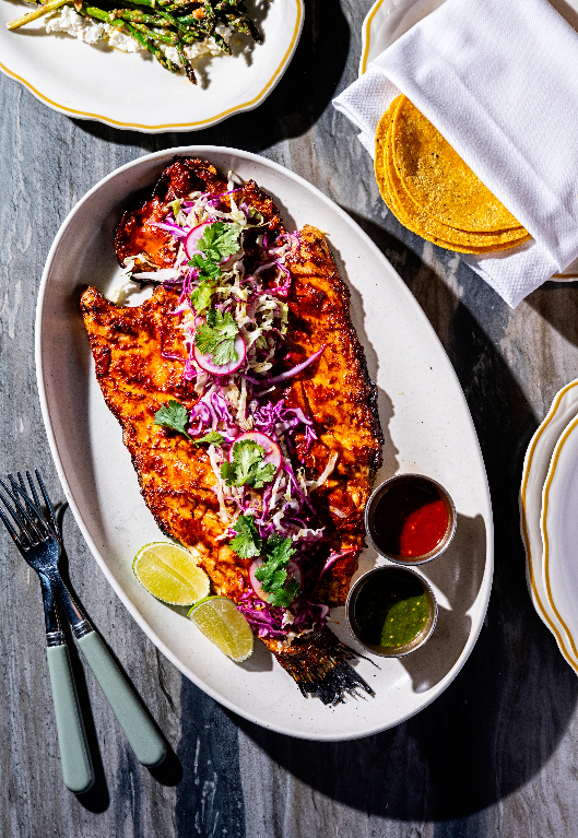 A whole grilled fish topped with red cabbage slaw and cilantro, served with lime wedges and dipping sauces.