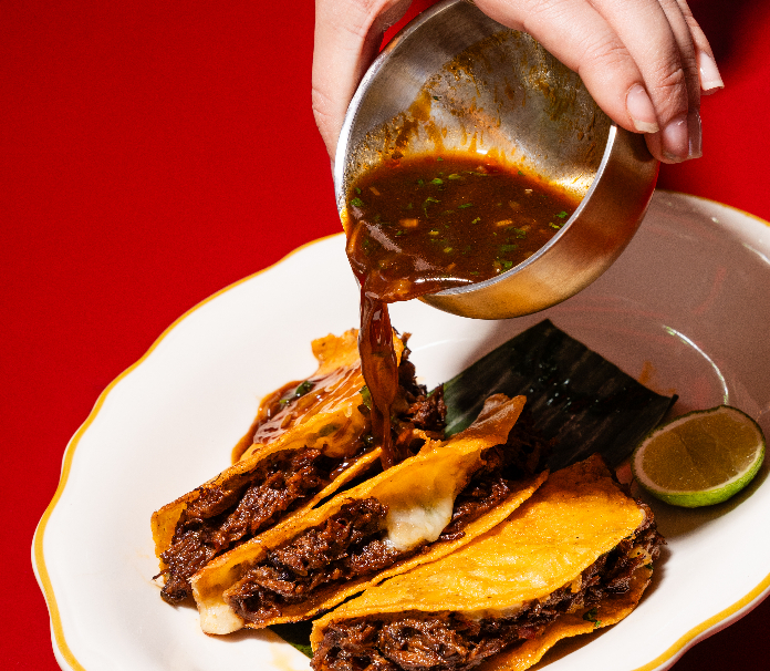 Rich broth being poured over Birria Tacos with melted cheese, served with a lime wedge on a red background.