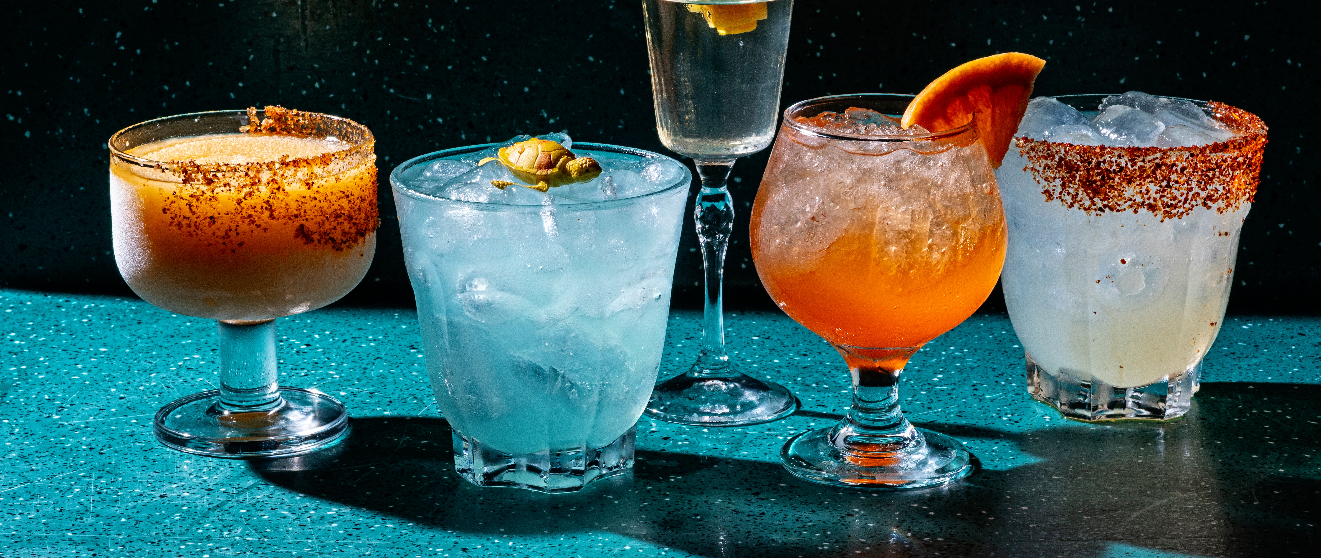 Five colorful margarita cocktails with salted rims, ice, and citrus garnishes arranged on a blue bar counter